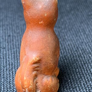 May include: A small, orange-brown ceramic cat figurine. The cat is sitting upright with its back to the viewer, showcasing its rounded form and textured surface. The ears are pointed, and the tail is tucked in. The figurine has a rustic, aged appearance.