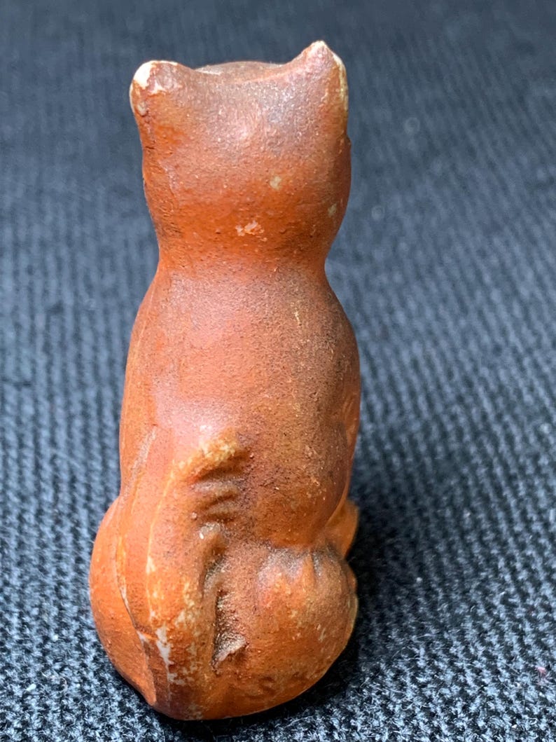 May include: A small, orange-brown ceramic cat figurine. The cat is sitting upright with its back to the viewer, showcasing its rounded form and textured surface. The ears are pointed, and the tail is tucked in. The figurine has a rustic, aged appearance.