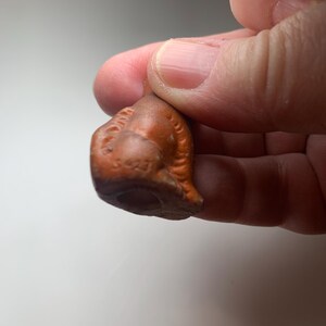 May include: A small, antique, brown ceramic figurine. The figurine has a detailed, textured surface with a rounded shape and a hole at the bottom. The number "3423" is visible on the front of the figurine.