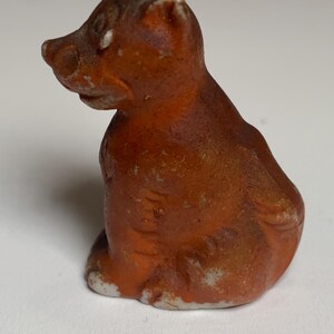 May include: A small, orange-brown ceramic figurine of a seated bear. The bear has a rounded body, small ears, and a pointed snout. The figurine has white paint on the base and around the edges, giving it a rustic appearance.