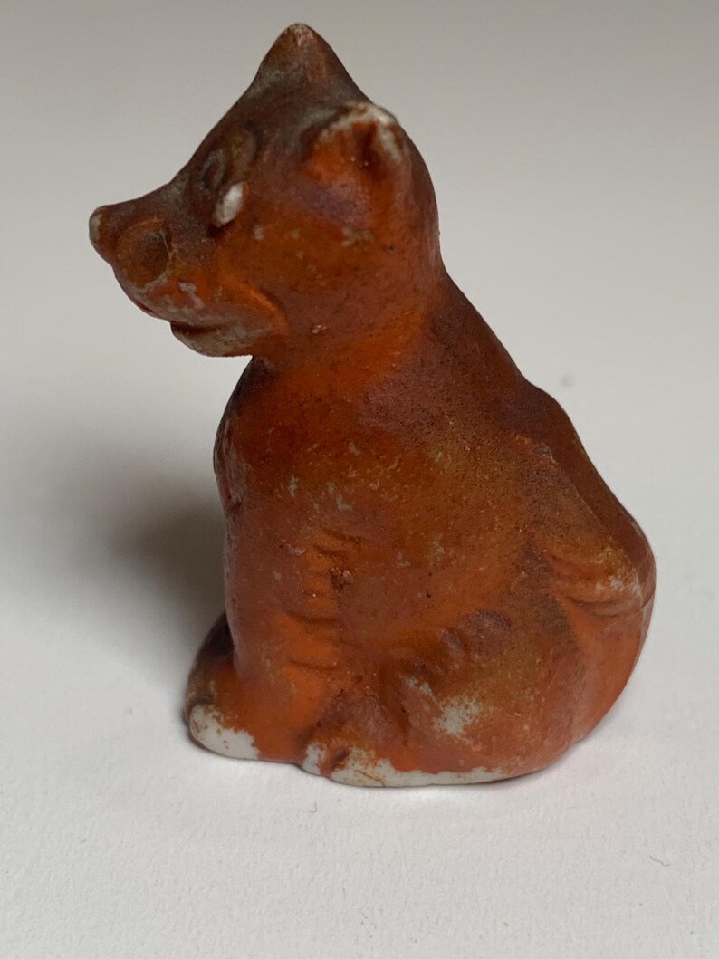 May include: A small, orange-brown ceramic figurine of a seated bear. The bear has a rounded body, small ears, and a pointed snout. The figurine has white paint on the base and around the edges, giving it a rustic appearance.