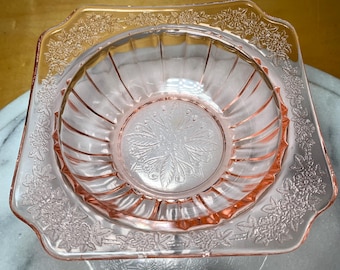 May include: Pink glass bowl with a square base and a round interior. The bowl has a floral design around the rim and in the center. The bowl is made of a transparent pink glass.