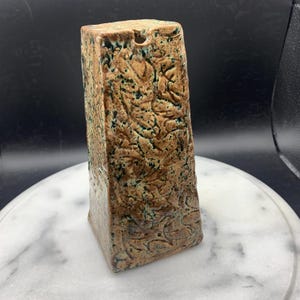 May include: A tall, four-sided ceramic vase with a tapered rectangular shape. The surface features a textured, mottled brown glaze with dark green speckles and deep, carved organic patterns throughout the exterior. A small circular opening is visible.