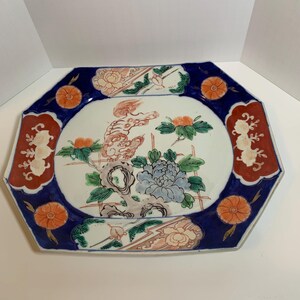 May include: An octagonal ceramic plate with a white center and a dark blue border. The plate features a floral design with orange, red, and blue accents. The center has a detailed illustration of a flower and a mythical creature.