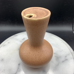 May include: A small, light brown ceramic vase with a speckled, matte finish. The vessel features a rounded, bulbous base that tapers upward into a slender, flared neck. The top opening is circular and slightly offset, showing a smooth, rounded rim.