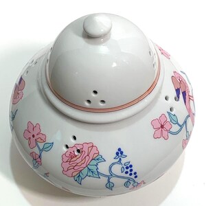May include: White ceramic teapot with a lid and floral design. The teapot features pink roses, blue accents, and a pink rim. The lid has a small knob and several small holes for steam release.