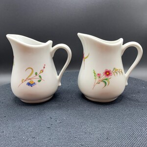 May include: Two white ceramic creamers with curved handles and pouring spouts. Each piece features a delicate hand-painted floral motif with green stems, yellow accents, and small blue or pink blossoms on the front surface of the rounded body.
