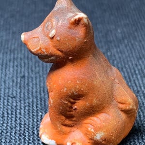 May include: A small, orange-brown ceramic figurine of a seated bear. The bear has a textured surface with white accents on the paws and ears. The figurine is a vintage style, with a slightly worn appearance.