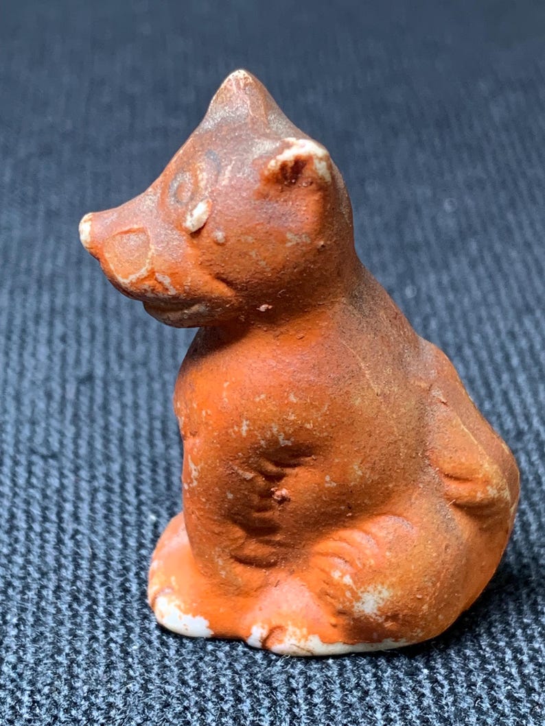 May include: A small, orange-brown ceramic figurine of a seated bear. The bear has a textured surface with white accents on the paws and ears. The figurine is a vintage style, with a slightly worn appearance.