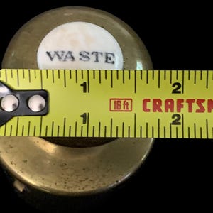 May include: A brass bottle stopper with a white porcelain top that says "WASTE" in black lettering. The stopper is measured with a yellow tape measure, showing it is about 1 inch in diameter.
