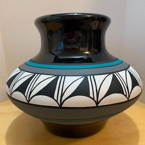 May include: A black and gray ceramic vase with a wide base and a flared rim. The vase features geometric patterns in white and black, with teal and gray bands. The glossy finish reflects light, highlighting the detailed design.
