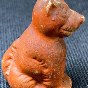 May include: A small, antique terracotta figurine of a seated cat. The cat is a reddish-brown color with visible texture and wear. The figurine has a detailed face and is posed in a sitting position, showcasing its small size and aged appearance.