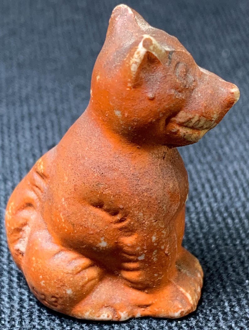 May include: A small, antique terracotta figurine of a seated cat. The cat is a reddish-brown color with visible texture and wear. The figurine has a detailed face and is posed in a sitting position, showcasing its small size and aged appearance.
