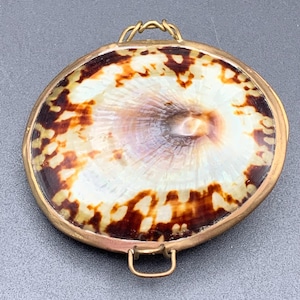 May include: A circular pendant featuring a polished, mottled brown and cream seashell inlay set within a smooth gold-toned metal frame. The piece includes two small, rectangular metal loops attached to the top and bottom edges of the frame.