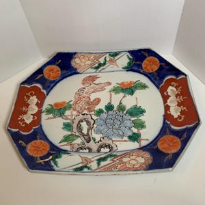 May include: An octagonal ceramic plate with a white center featuring a floral design with blue, orange, and green accents. The border is dark blue with orange floral patterns and red accents.