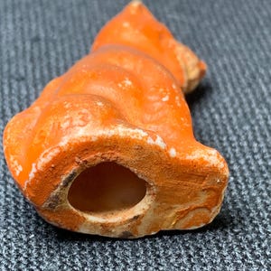 May include: A small, orange ceramic cat figurine with a hollow base. The cat is in a seated position, with visible details including its ears and tail. The figurine has a distressed, aged appearance with white and brown accents. The number "23" is visible on the side.