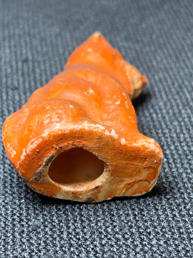 May include: A small, orange ceramic cat figurine with a hollow base. The cat is in a seated position, with visible details including its ears and tail. The figurine has a distressed, aged appearance with white and brown accents. The number "23" is visible on the side.