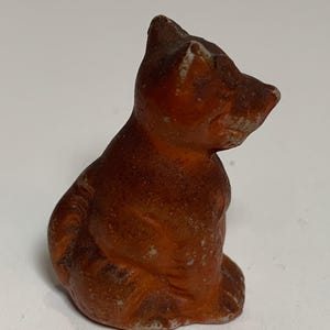 May include: A small, brown figurine of a seated cat. The cat is in a sitting position, with its head turned to the side. The figurine has a weathered appearance, with visible wear and tear. The cat's fur is a mix of brown and orange hues.