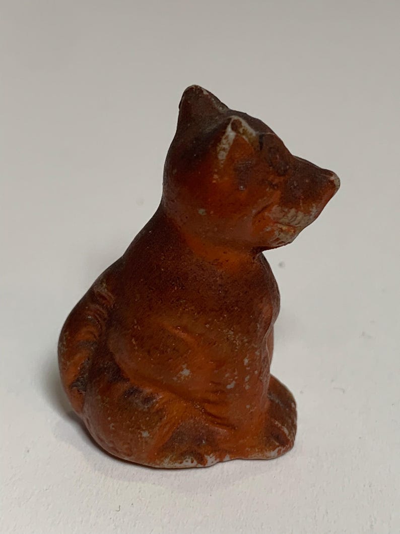 May include: A small, brown figurine of a seated cat. The cat is in a sitting position, with its head turned to the side. The figurine has a weathered appearance, with visible wear and tear. The cat's fur is a mix of brown and orange hues.