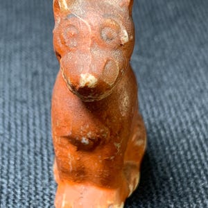 May include: A small, antique-style figurine of a seated cat. The cat is primarily orange-brown with lighter accents on the face and paws. The figurine has a weathered appearance, suggesting age or intentional distressing.