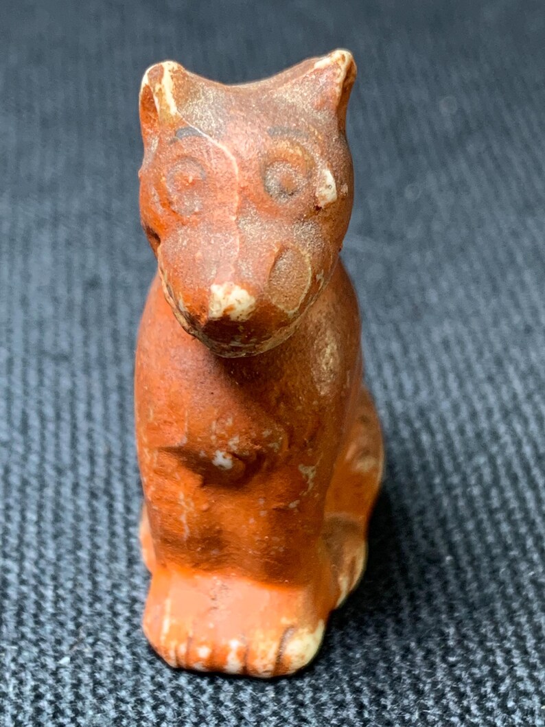May include: A small, antique-style figurine of a seated cat. The cat is primarily orange-brown with lighter accents on the face and paws. The figurine has a weathered appearance, suggesting age or intentional distressing.