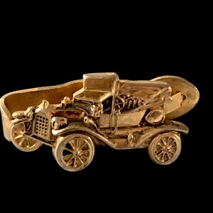 May include: Gold-toned metal tie clip featuring a detailed model of a vintage car. The car has a black grill and a black steering wheel.