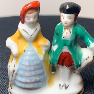 May include: A small, vintage porcelain figurine depicting a couple in period attire. The woman wears a yellow coat with a red hat, while the man has a green jacket and black hat. The base is white with gold accents.