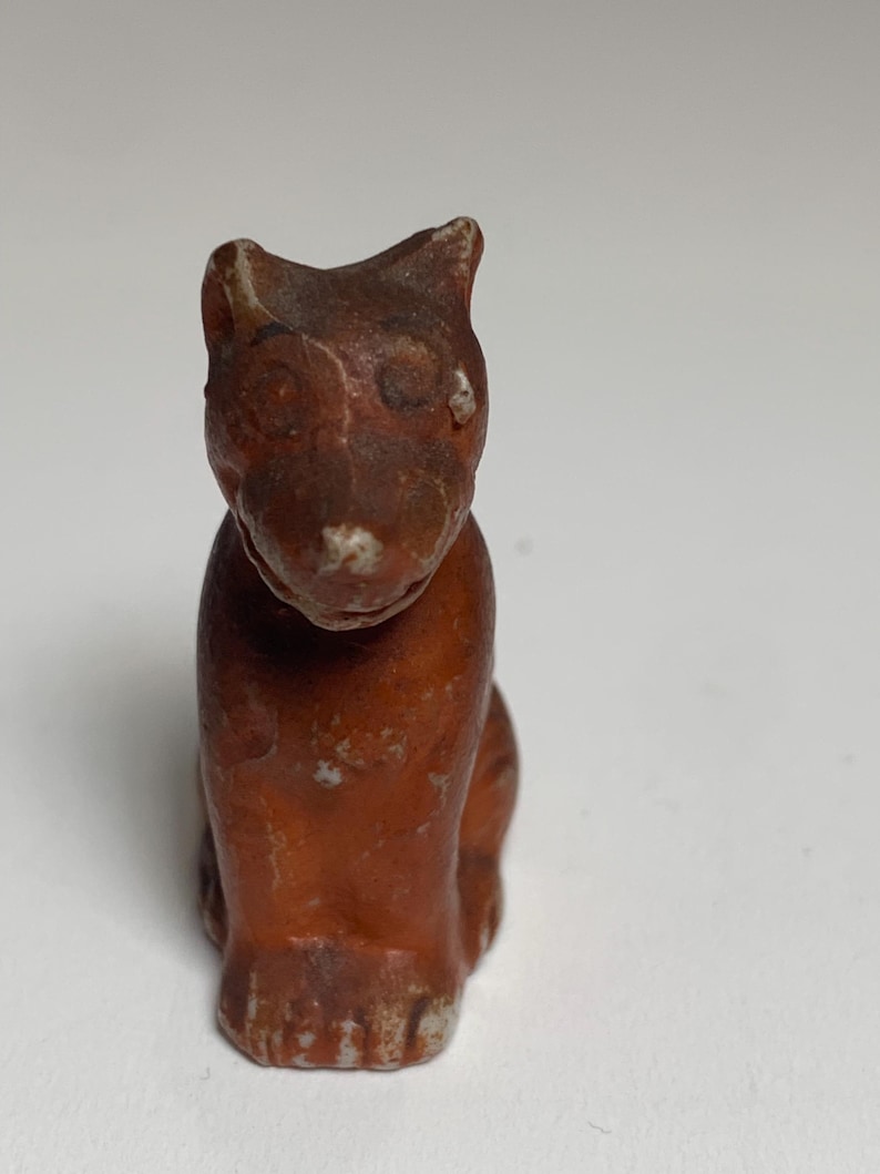 May include: A small, brown, cat figurine with a sitting posture. The figurine has a weathered appearance with visible wear and tear, and a light-colored patch on its chest. The cat's ears are pointed, and its eyes are visible.