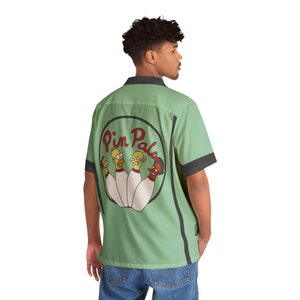 May include: A green bowling shirt with black trim and a circular patch on the back. The patch features a cartoon image of four bowling pins with faces, and the text "Pin Pals".
