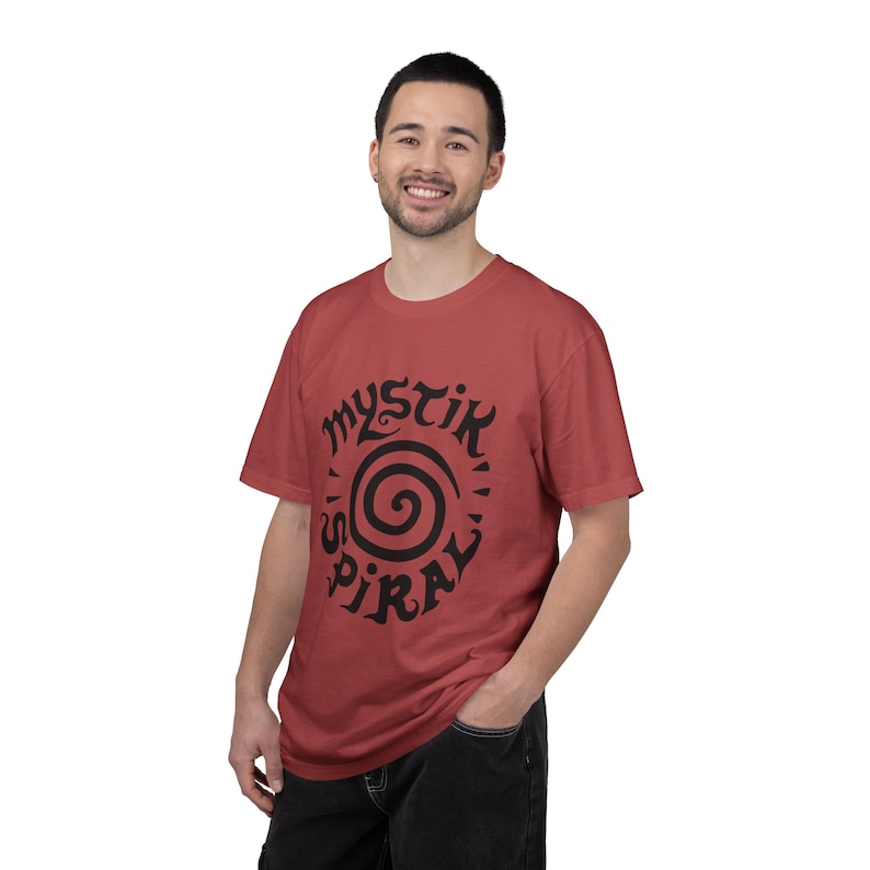 K&ouml;nnte beinhalten: Ein ziegelrotes Kurzarm-T-Shirt mit einem schwarzen Grafikdruck auf der Brust. Das Design besteht aus einer zentralen Spirale, umgeben von dem Schriftzug "MYSTIK SPIRAL" in einer stilisierten, fetten, dunklen Schrift. Das Shirt hat einen Rundhalsausschnitt.