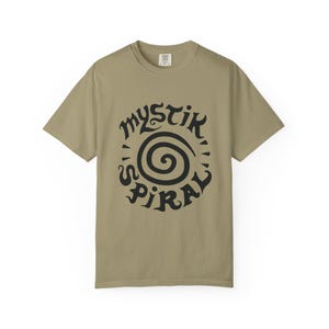 K&ouml;nnte beinhalten: Ein khakifarbenes Kurzarm-T-Shirt mit einem schwarzen Grafikdruck auf der Brust. Das Design besteht aus einer zentralen Spirale, die von dem Text "MYSTIK SPIRAL" in einer stilisierten, fetten und dunklen Schriftart umgeben ist.