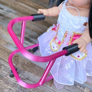 Puede incluir: Un andador de metal rosa con asas y ruedas negras. El andador está siendo utilizado por una muñeca que lleva un vestido blanco con detalles en rosa y dorado.