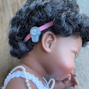 May include: A close-up of a baby doll with dark curly hair wearing a white dress with a pearl necklace. The doll has a hearing aid behind her ear with a pink strap.