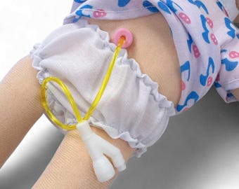 Könnte beinhalten: Eine Puppe mit einem hellblau-weiß gemusterten Oberteil und weißen Rüschenshorts. Ein gelber Schlauch und ein weißer Y-förmiger Stecker sind am Bein der Puppe befestigt, mit einem rosa kreisförmigen Teil am Körper der Puppe.