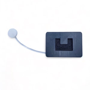 May include: A black plastic device with a small white button on a cord. The device has a rectangular shape with a cutout in the center.