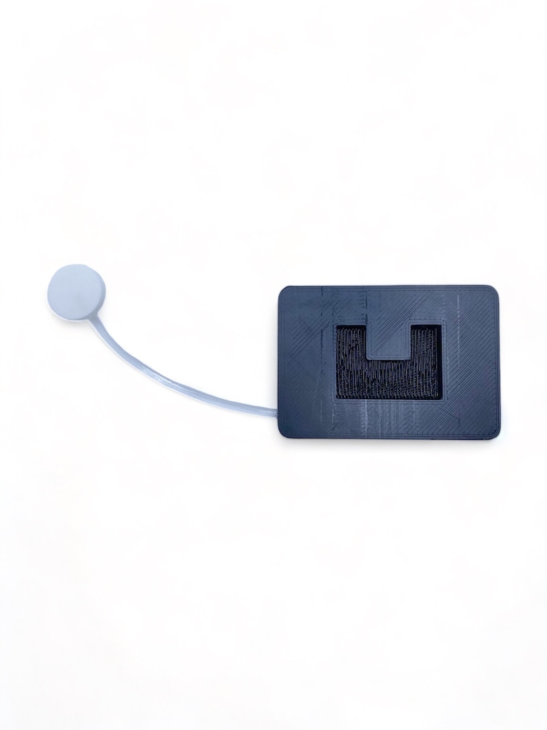 May include: A black plastic device with a small white button on a cord. The device has a rectangular shape with a cutout in the center.