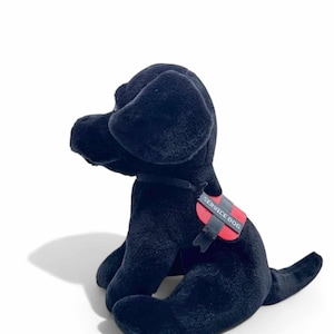 May include: A plush black Labrador Retriever service dog toy, seated with its head turned to the left. It wears a red and black service dog vest with the words "SERVICE DOG" printed on it.