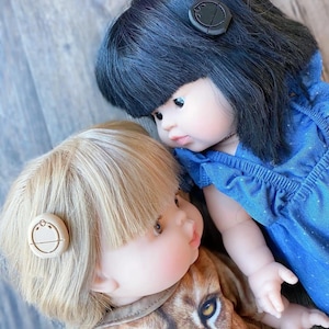 May include: Two dolls with different hair colors and outfits. The doll on the left has blonde hair and is wearing a brown and orange shirt with a lion print. The doll on the right has black hair and is wearing a blue dress. Both dolls have a round, wooden button on their hair.