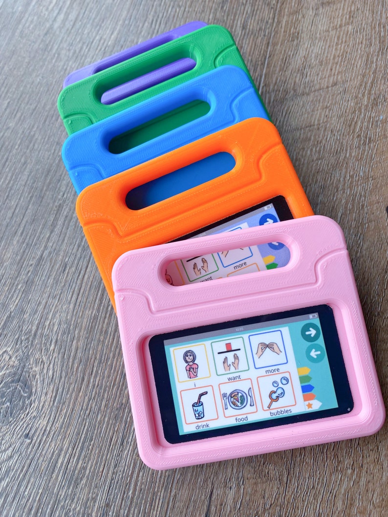 May include: A stack of four rectangular tablet cases in pink, orange, blue, and green, each featuring a built-in handle. The front-most pink case holds a tablet displaying a communication grid with icons labeled i, want, more, drink, food, and bubbles.