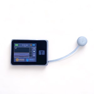 May include: A black and gray medical device with a small screen displaying a graph and the text "11:14 AM" and "112 mg/dL". The device has a blue button labeled "BOLUS" and an orange button labeled "OPTIONS". A gray tube with a round gray end is attached to the device.