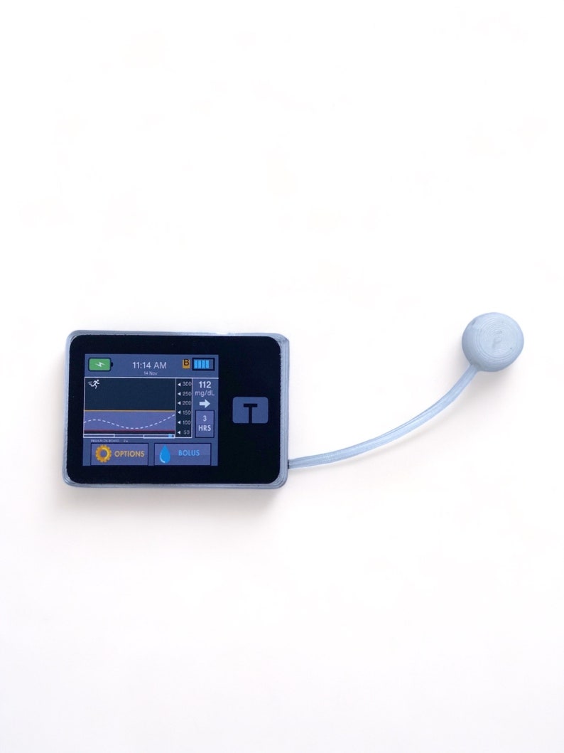 May include: A black and gray medical device with a small screen displaying a graph and the text "11:14 AM" and "112 mg/dL". The device has a blue button labeled "BOLUS" and an orange button labeled "OPTIONS". A gray tube with a round gray end is attached to the device.