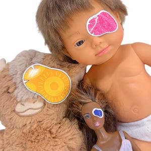 Op de afbeelding: Een verzameling speelgoed, waaronder een pop, een pluche koe en een pop met bruin haar. Elk speeltje heeft een sticker over één oog. De stickers zijn roze, geel en blauw.