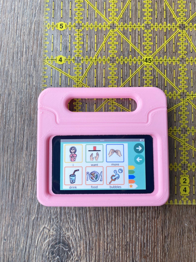 May include: A pink rectangular communication device case with a built-in handle, featuring a screen displaying six icons labeled i, want, more, drink, food, and bubbles. A yellow quilting ruler is positioned behind the case for scale.