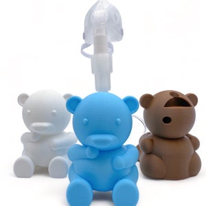 May include: Three teddy bear-shaped nebulizer holders in white, blue, and brown. A clear plastic nebulizer mask is on top of the blue bear.