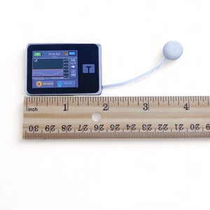 May include: A small, gray, rectangular medical device with a black screen displaying a graph and the time 11:14 AM. The device has two buttons labeled "OPTIONS" and "BOLUS". A white tube with a round, gray sensor is attached to the device.