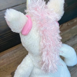 May include: A white plush unicorn with a pink mane and a pink ear tag. The unicorn has a sparkly horn.