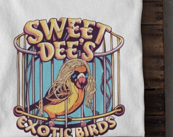May include: White t-shirt with a graphic of a yellow bird with long blonde hair and sunglasses in a cage. The text "Sweet Dee's Exotic Birds" is printed below the cage.