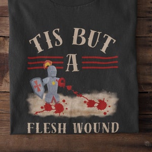 May include: Black t-shirt with the phrase "TIS BUT A FLESH WOUND" in a vintage font. The design includes a cartoon knight with a shield and a sword, with red blood splatter effects.