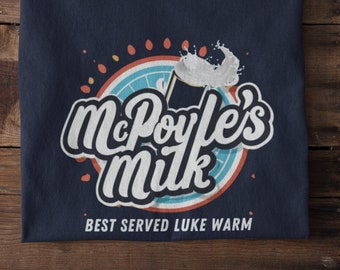May include: Navy blue t-shirt featuring the text "McPoyle's Milk" in a stylized font, with a milk splash graphic. The design includes a circular element with orange and blue accents, and the words "BEST SERVED LUKE WARM" are printed below.