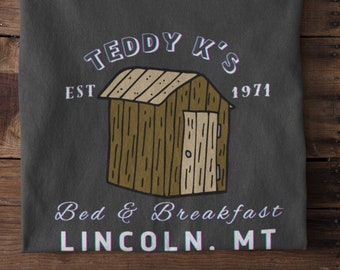 May include: A dark gray t-shirt with a vintage-style graphic design. The design features a small wooden cabin with a door and a roof. The text "Teddy K's" is above the cabin, and the text "Est 1971" is to the right of the cabin. The text "Bed & Breakfast" is below the cabin, and the text "Lincoln, MT" is below that.